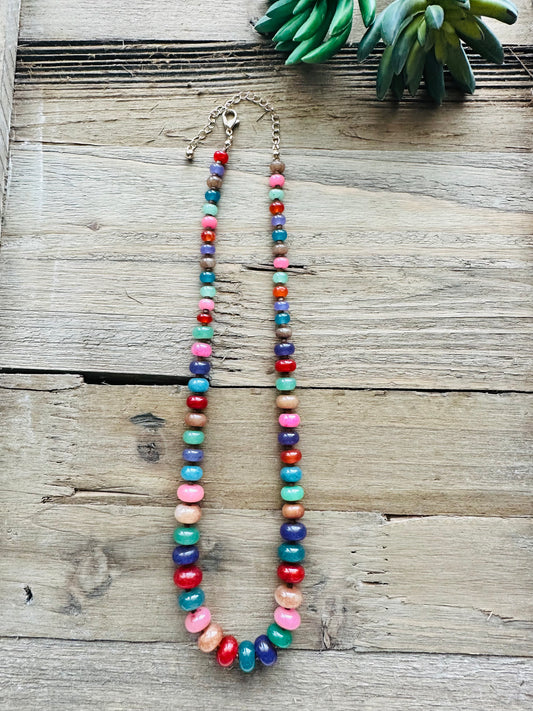 A colorful beaded necklace with alternating round beads in red, pink, teal, blue, green, and brown, displayed on a rustic wooden surface next to a green succulent.