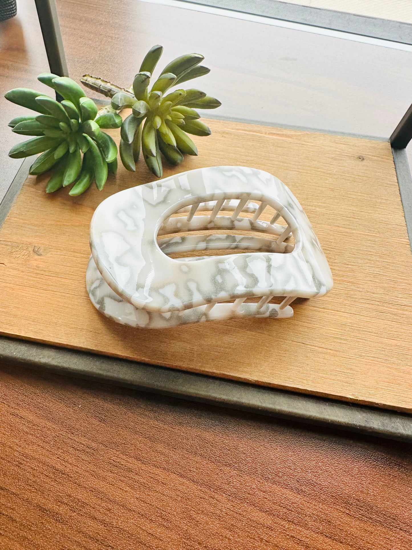 A white and gray marbled claw hair clip styled on a wooden tray next to a small green succulent plant.