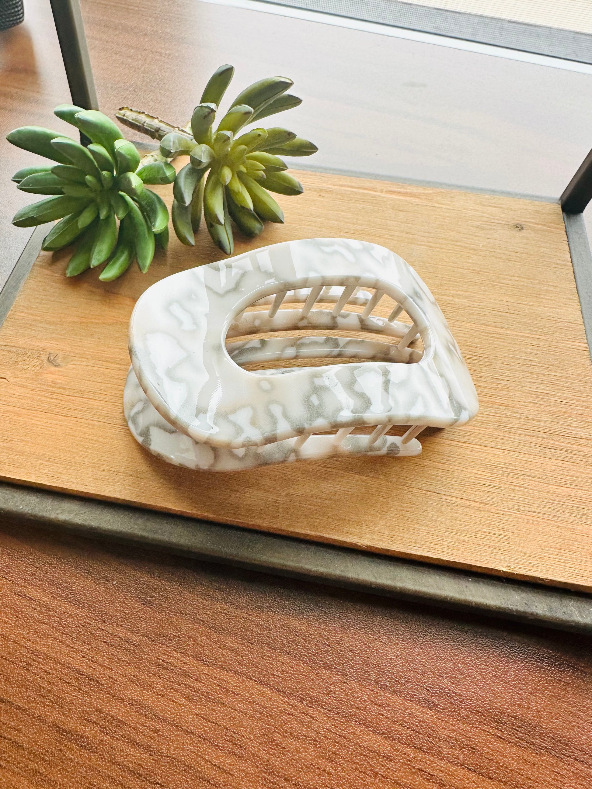 A white and gray marbled claw hair clip styled on a wooden tray next to a small green succulent plant.