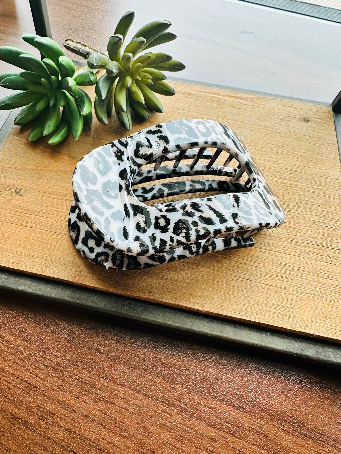 A hair claw clip with a black and white leopard print fabric, resting on a wooden cutting board next to a small green succulent plant.