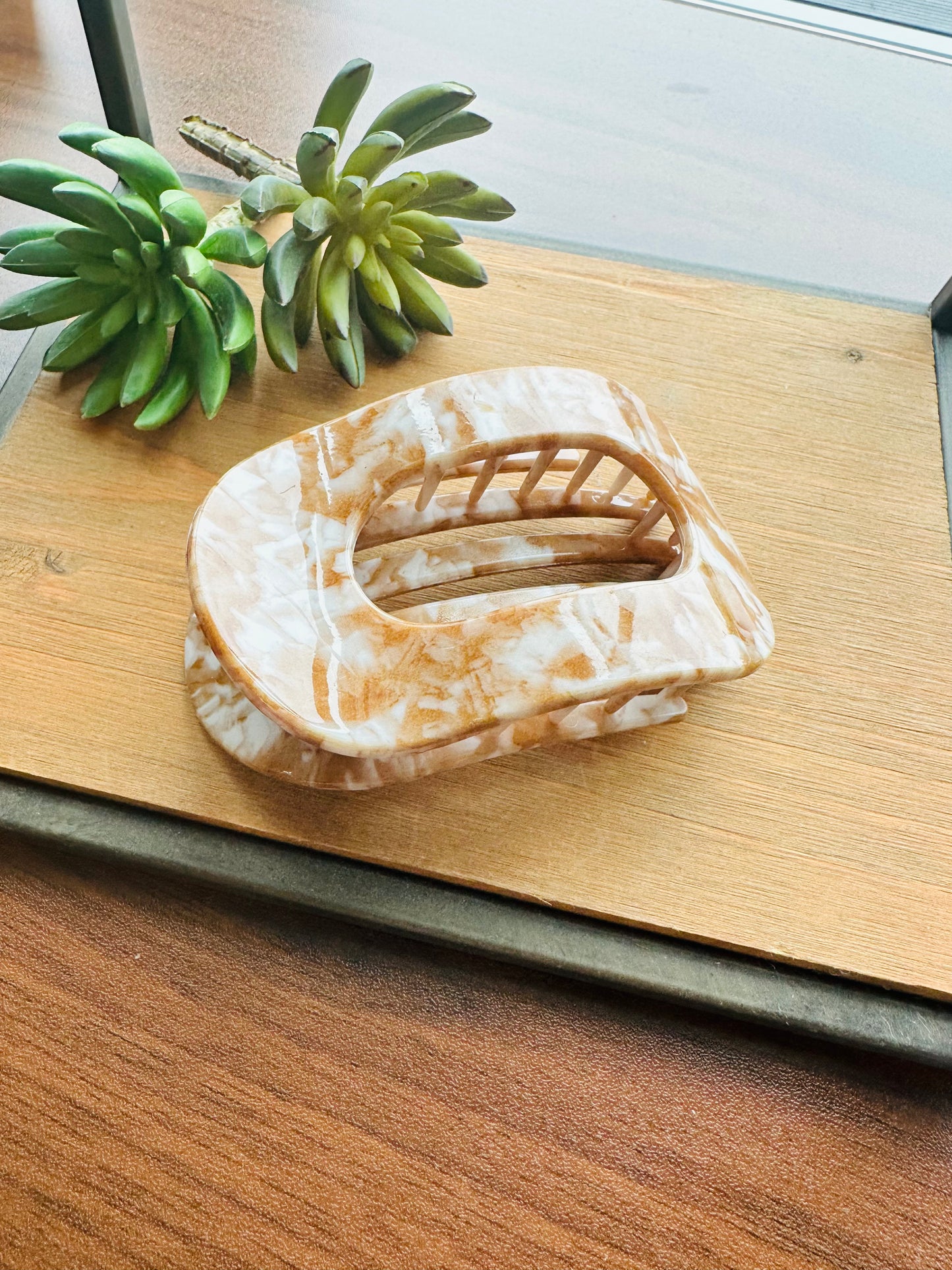 A tortoiseshell hair claw clip with a marbled beige and brown pattern, displayed on a wooden tray with small green succulent plants.