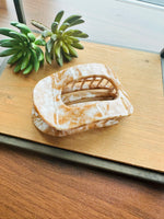 A tortoiseshell hair claw clip with a marbled beige and brown pattern, displayed on a wooden tray with small green succulent plants.