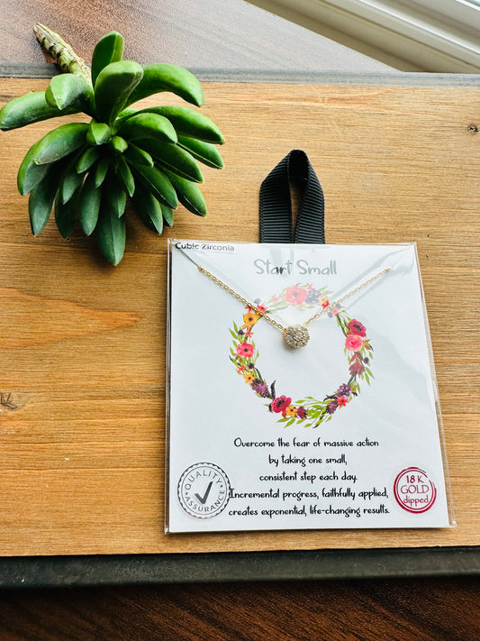 A gold necklace with a small floral wreath pendant is displayed on its white retail card, which hangs from a black ribbon. The card is placed on a wooden surface next to a green succulent plant.