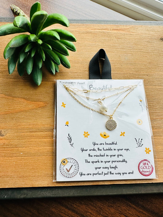 A gold double-layer necklace with a white pearl and round gold pendant, presented on a white card with decorative text and illustrations, resting on a wooden table next to a green succulent.